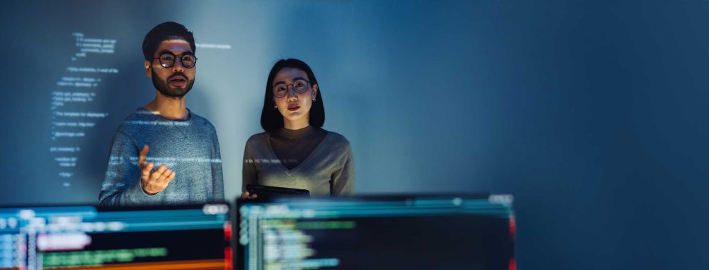 Asian Two software developers engaged in collaborative coding session, with code projected onto the wall and multiple computer screens displaying